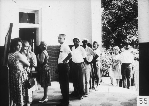 A line of people stand outdoors in front of the open doorway of a building. Included are both Caucasian and African-American men and women.