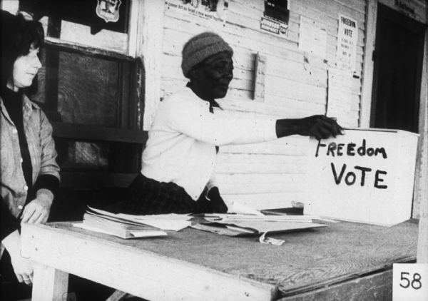 Two women sit behind a table at a Freedom Vote, a mock election designed to include African-Americans who were unable to register to vote and would be a truer reflection of how Mississippi would vote. Freedom Votes were held on October 30 and 31, and November 1 and 2.  A box on the table has the words: "Freedom Vote" written on it.