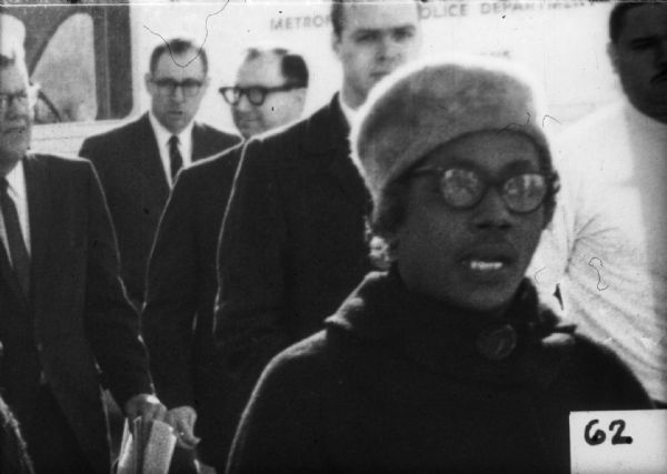 A group of people is led by Annie Devine wearing a hat and eyeglasses in the foreground, along with several men in the background. Behind them is a vehicle with a sign that says, in part, "Metrop[...] [...]olice Depart[...]".
