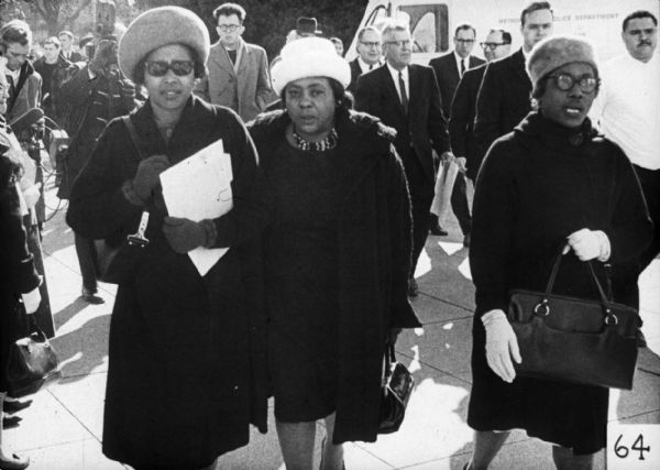 Annie Devine, Fannie Lou Hamer, and Victoria Gray, fellow MFDP candidates. All three women are wearing hats and carrying handbags. A crowd of people are behind them, including someone using a movie or television camera, and someone else holding a large microphone. Also in the background is a truck on which is written: Metrop[...] [...]olice Department."