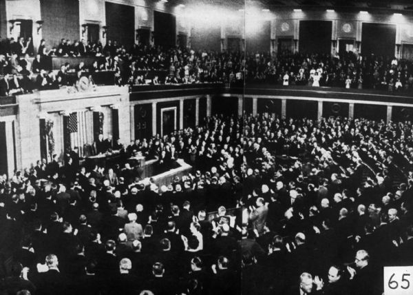 An elevated view of a crowded room. People are in the balcony and also on the main floor, surrounding a group of men on a raised platform in front of a flag. The crowd is standing and they all appear to be applauding.