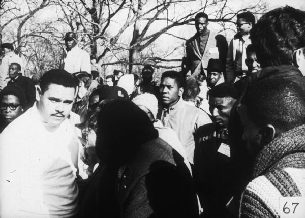 An integrated group of people stand outdoors in coats and hats. A man in a light-colored shirt in the foreground, probably civil rights activist and MFDP member Lawrence Guyot, looks into the camera.