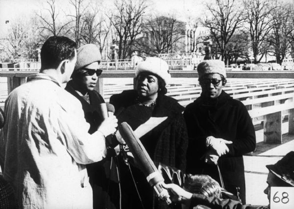 Fannie Lou Hamer stands outdoors along with Annie Devine and Victoria Gray, being interviewed by a man with a microphone. At the bottom of the frame, two other men are crouching and holding microphones. In the background is a large building with columns.
