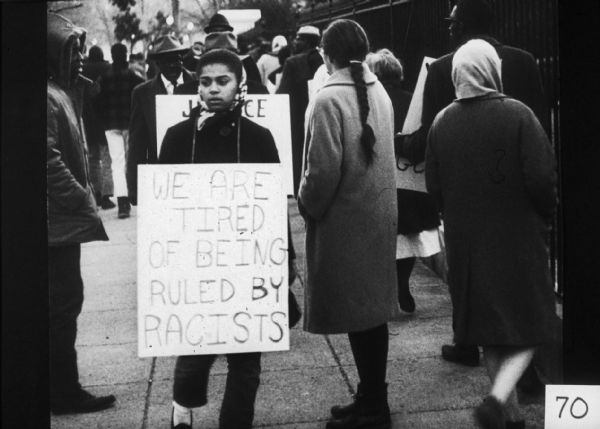 A girl at a demonstration holding a sign that reads: "We are tired of being ruled by racists." Behind her, other people carry signs as they walk along a fence.