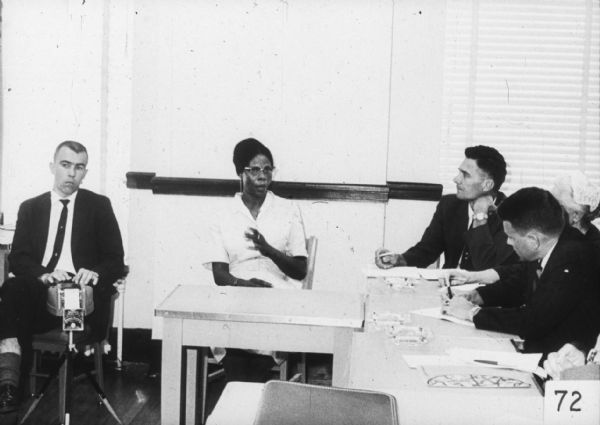 An African-American woman is seated behind a small table while speaking. On the left a stenographer types, and on the right a group of people sit and listen.