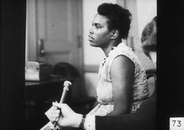 An African-American woman sits in a chair, while a man in the foreground holds a microphone for her to speaking in to.