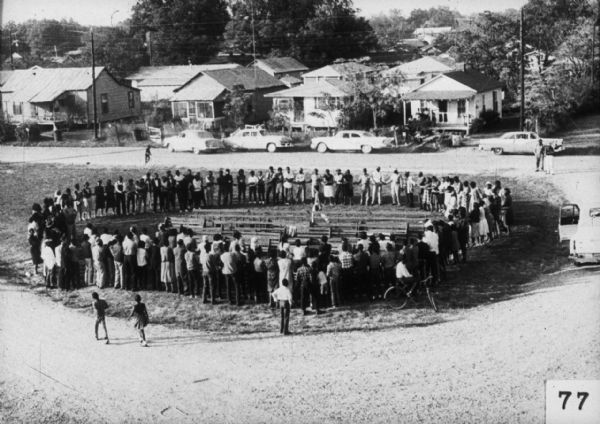 An elevated view of a large group of people standing in a circle around empty benches holding hands. Vehicles are parked near houses in the background.