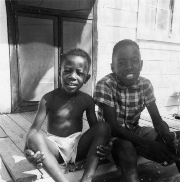 Two boys on a porch pose for a civil rights volunteer. "Tyrone (right) and his brother — my first friends whose parents allowed me to use their large bed during my over-night stay to Greenville. Prior to this picture, we played football and Tyrone gave me my first piece of okra from his garden."