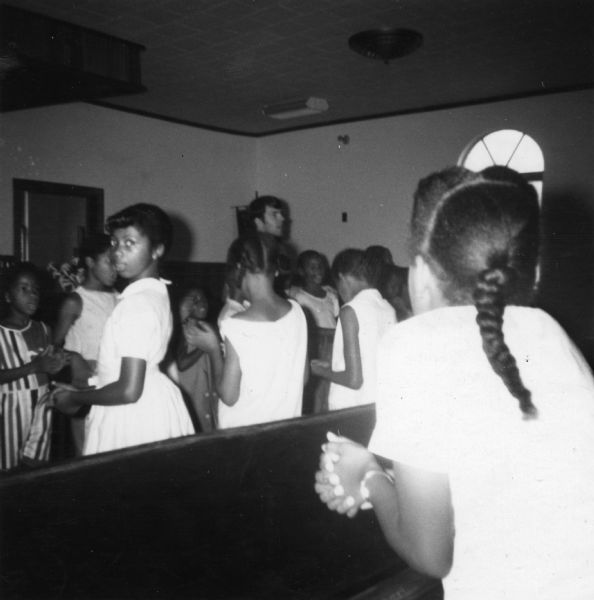 Students in a Freedom School singing and clapping in an image taken by a civil rights volunteer.

"Freedom School - Freedom singing led by youngsters and Dennis Selig, a young man from Chicago who was the obvious favorite teacher in the Freedom School.  Occasionally he would leave the school in order to demonstrate, e.g., he formed the picket line of the Holiday Sun strike of the seven maids."