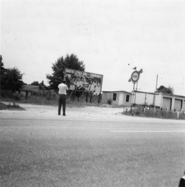 Image of a billboard near a road taken by a civil rights volunteer.  A man stands in front of the billboard across the road.<p>"'Martin Luther King at Communist Training School' reads and pictures the billboard along Mississippi highways. Personally, I thought how wonderful it was to see a friend's picture on the Mississippi highways every now and then."