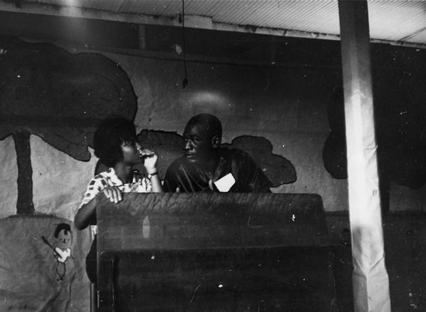 A man and a woman speak at a podium in front of a mural at the Freedom School convention during Freedom Summer.