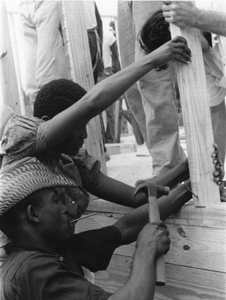 Three people working on a community construction project. All three support a beam, while the man in the foreground hammers a nail into it.