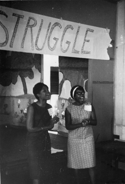 Two women clap and sing at the Freedom School Convention during the Freedom Summer civil rights project. Behind them is a mural, and a banner that says "Struggle" hangs above them.