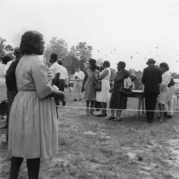 A line of predominantly African-American men and women waiting in a line at a primary election for the LCFO (Lowndes County Freedom Organization). In the background, a Caucasian man holding a camera stands behind the line.