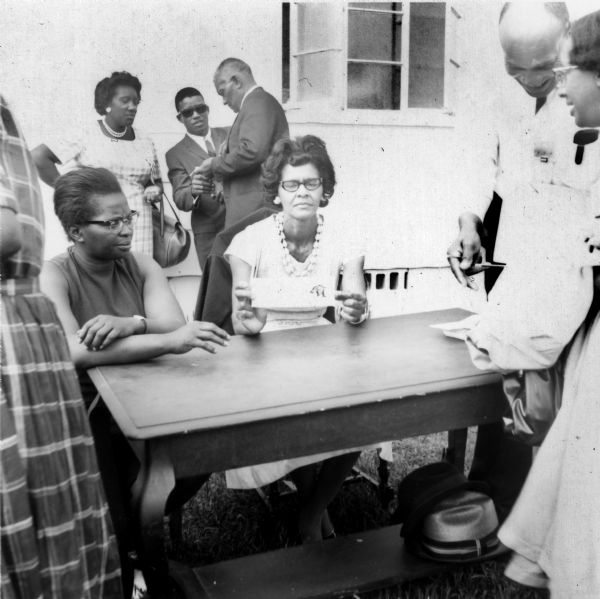 Two women are outdoors seated at a table in front of a building, surrounded by several people. One of the women is holding a document in her hands, showing a black panther. The black panther was the logo for the LCFO (Lowndes County Freedom Organization), a civil rights group that directly influenced the later Black Panther Party.
