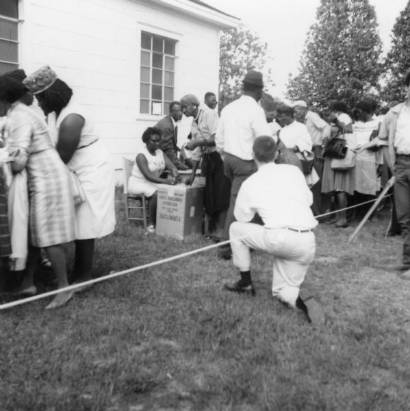 A line of people behind a rope wait near tables outdoors near a building at a primary election for the LCFO (Lowndes County Freedom Organization). In the foreground, a man kneels near the rope with his back to the camera, probably taking photographs.