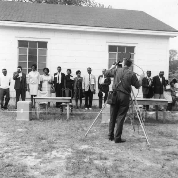 A group of African-American men and women, probably voter registration volunteers, stand in a line outdoors in front of a building at a primary election for the LCFO (Lowndes County Freedom Organization). In front of them are tables and chairs. In the foreground, a Caucasian man operates film recording equipment.