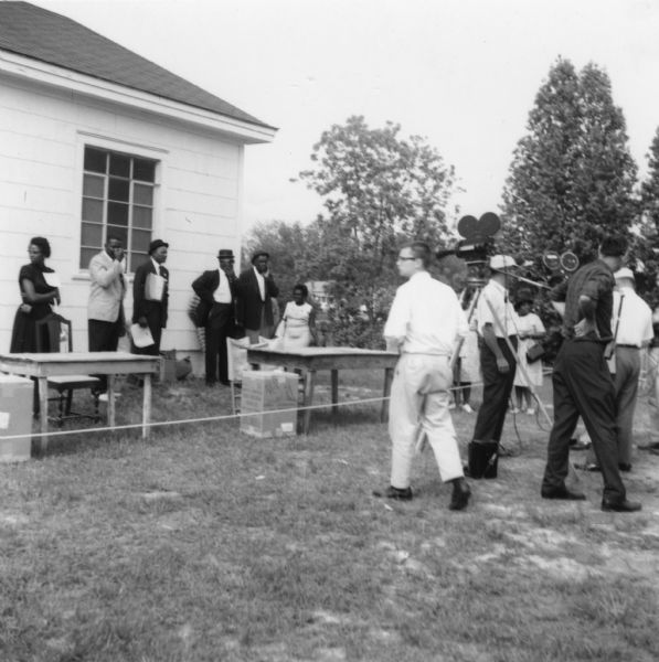 Several people, probably voter registration volunteers, stand outdoors in front of a building near tables and chairs at a primary election for the LCFO (Lowndes County Freedom Organization). Nearby, a group of men with film equipment work.