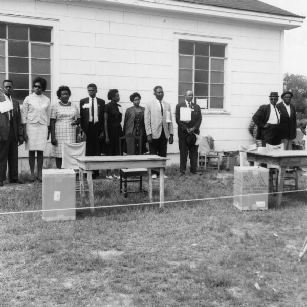 Several people, probably voter registration volunteers, stand outdoors next in front of a building near tables and chairs.  They are taking part in a primary election for the LCFO (Lowndes County Freedom Organization).