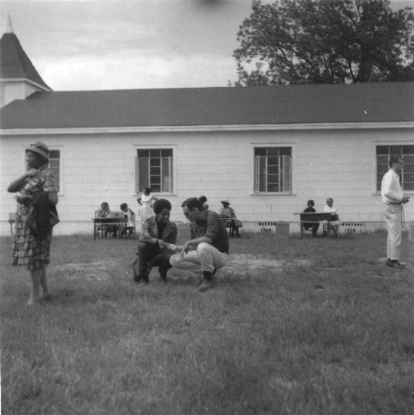 Several people stand outdoors on a lawn near a building at a primary election for the Lowndes County Freedom Organization (LCFO). In the center, an African-American woman and a Caucasian man crouch near the ground, talking.