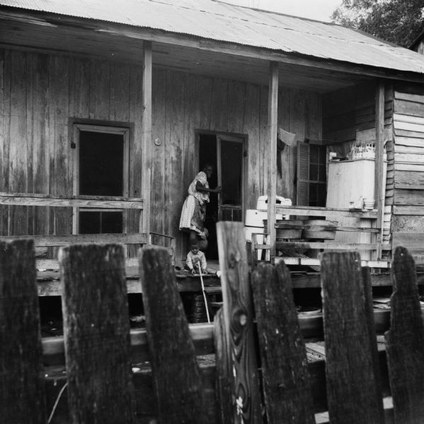 A small child is seated on a porch while a woman stands in the doorway. "Yards turn to mudd [sic] when it rains — children must play on broken and rotting porches."<p>Beekman Quarters housed 81 people in 23 buildings. None of the houses in the neighborhood had running water or gas/electric heating; the neighborhood had 15 outdoor toilets and 11 outdoor water hydrants.