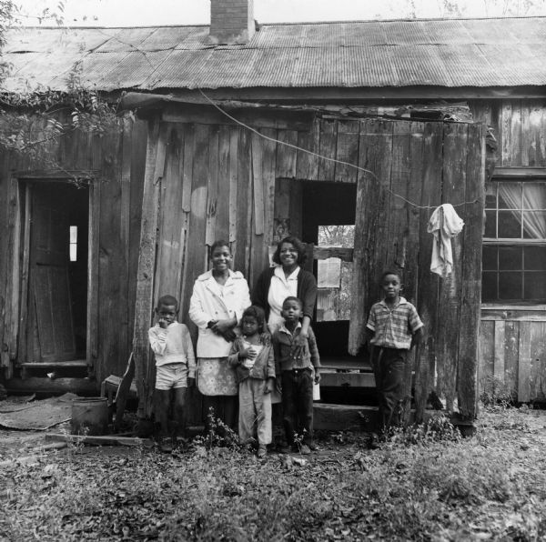 A family poses in front of their home. "This family has to stop using the back room to this house. What used to be a doorway is now part of a huge hole in the wall. Father earns $30.00 a week on a full-time job."<p>Beekman Quarters housed 81 people in 23 buildings. None of the houses in the neighborhood had running water or gas/electric heating; the neighborhood had 15 outdoor toilets and 11 outdoor water hydrants.