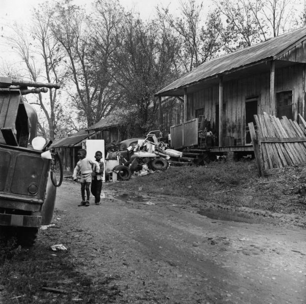 Two boys stand in a wet dirt road in the foreground. In the background is a house with a pile of objects in the yard, including an automobile, a door, and tires. "One tenant collects junk and neither he nor landlord will oblige other tenants by removing it."<p>Beekman Quarters housed 81 people in 23 buildings. None of the houses in the neighborhood had running water or gas/electric heating; the neighborhood had 15 outdoor toilets and 11 outdoor water hydrants.
