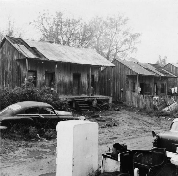 Several houses along a dirt road. A person stands in the doorway of one house. "'Some roofs leak worse than others, but we all get wet inside when it rains.' 'And when it's cold the wind goes right on through.' 'Sure we asked him to fix it, but it don't do no good.'"<p>Beekman Quarters housed 81 people in 23 buildings. None of the houses in the neighborhood had running water or gas/electric heating; the neighborhood had 15 outdoor toilets and 11 outdoor water hydrants.