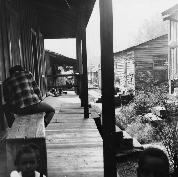 Porches line a walkway. Two children and a man sitting on a bench are in the foreground, while a dog and a child are visible in the background.<p>“Row upon row of dilapidated buildings, three rows deep, characterize this slum. The city has no housing code and continues to ignore the slums of Natchez. No pavement, no streetlights, 10 outside flush toilets in 2 centrally located shanties shared by 114 people. The 125 persons living in this area share eight water faucets; of these 8, two are inside (installed by tenants) and 6 are outside.”