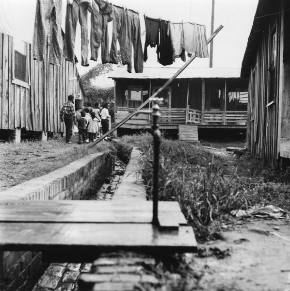 A water hydrant stands next to a drainage ditch, while clothing hangs on a clothes like above. In the background, children walk away from the camera between two houses. "This hydrant (also in pictures B-15, B-12) is the sole supply of water to 50 people in 12 families."<p>“Row upon row of dilapidated buildings, three rows deep, characterize this slum. The city has no housing code and continues to ignore the slums of Natchez. No pavement, no streetlights, 10 outside flush toilets in 2 centrally located shanties shared by 114 people. The 125 persons living in this area share eight water faucets; of these 8, two are inside (installed by tenants) and 6 are outside.”
