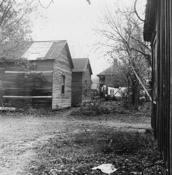 Buildings, likely houses, at the intersection of two dirt roads.  Laundry hangs from a clothesline in the background.<p>“Row upon row of dilapidated buildings, three rows deep, characterize this slum. The city has no housing code and continues to ignore the slums of Natchez. No pavement, no streetlights, 10 outside flush toilets in 2 centrally located shanties shared by 114 people. The 125 persons living in this area share eight water faucets; of these 8, two are inside (installed by tenants) and 6 are outside.”
