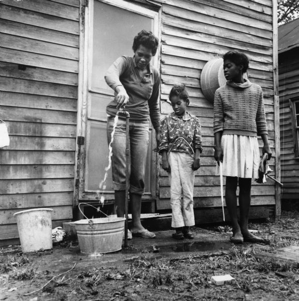 A woman and two girls look on as a water hydrant is turned on by the woman. "Two families, 6 people, share this hydrant."<p>“Row upon row of dilapidated buildings, three rows deep, characterize this slum. The city has no housing code and continues to ignore the slums of Natchez. No pavement, no streetlights, 10 outside flush toilets in 2 centrally located shanties shared by 114 people. The 125 persons living in this area share eight water faucets; of these 8, two are inside (installed by tenants) and 6 are outside.”