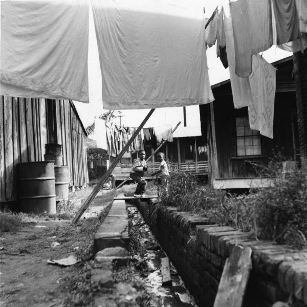 Two children look towards the photographer by a drainage ditch with buildings on all sides. Laundry hangs from clotheslines above.<p>“Row upon row of dilapidated buildings, three rows deep, characterize this slum. The city has no housing code and continues to ignore the slums of Natchez. No pavement, no streetlights, 10 outside flush toilets in 2 centrally located shanties shared by 114 people. The 125 persons living in this area share eight water faucets; of these 8, two are inside (installed by tenants) and 6 are outside.”
