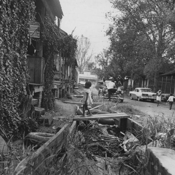 A child walks across ditch on a plank walkway. On the street, children play with balloons while three men recline on the hood of a car.<p>“Row upon row of dilapidated buildings, three rows deep, characterize this slum. The city has no housing code and continues to ignore the slums of Natchez. No pavement, no streetlights, 10 outside flush toilets in 2 centrally located shanties shared by 114 people. The 125 persons living in this area share eight water faucets; of these 8, two are inside (installed by tenants) and 6 are outside.”