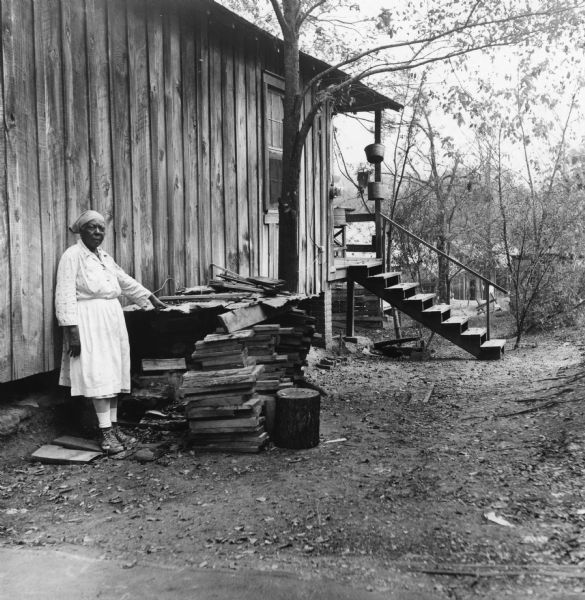 A woman stands next to a home near a woodpile. Behind the trees in the background, more homes are visible. "Houses are built in rows and cover the hill from the top down to the valley."<p>80 people live in 21 buildings in the Taylor Alley neighborhood of Natchez. The occupants have 11 hydrants, two indoors, and 15 toilets.