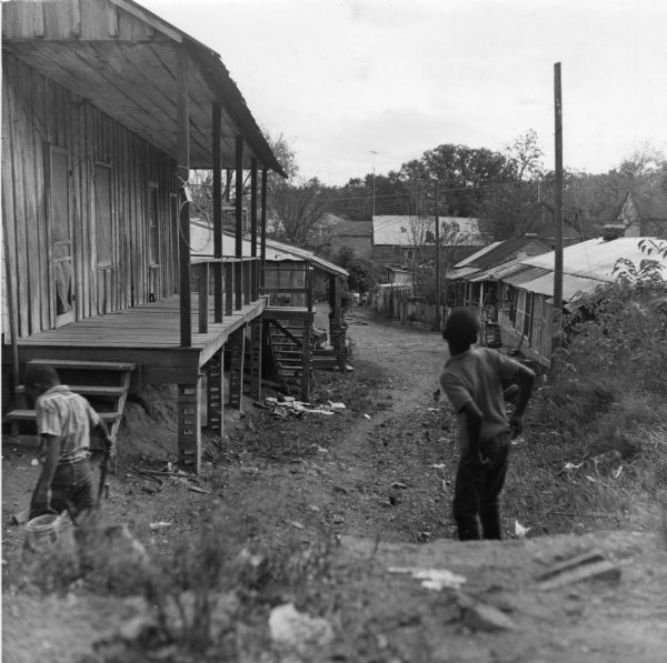 "A water faucet located at the top of the hill (the upper end of the alley) behind the boy is used by eleven families, 28 people, who live in the buildings below. It's a long walk." View down alley, with two children, and a chicken, in the foreground. In the background two women, obscured by columns, sit on a porch, and on the other side of the alley a man sits on another porch.<p>The De Marco Alley neighborhood houses 73 people in 18 buildings connected with dirt roads. Tenants share 6 outhouses and 1 outdoor flush toilet, as well as 10 water hydrants (1 indoors).