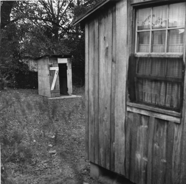 "Twelve families, eighteen persons, share four outhouses." A window of a house is visible in the foreground, while an outhouse is located behind the home.<p>Griffin Alley houses 12 families in 6 buildings. All residents share 2 hydrants and 4 toilets.