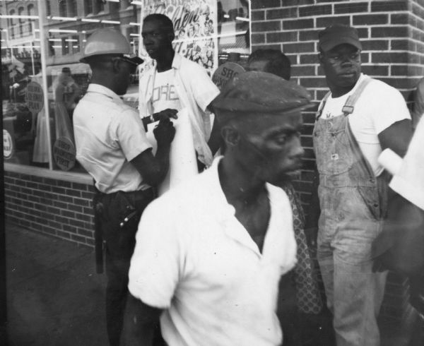 A protest of Woolworth's department store during Freedom Summer. A line of people are standing on the sidewalk in front of Woolworth's show windows while a police officer wearing a helmet writes on a large sheet of paper.