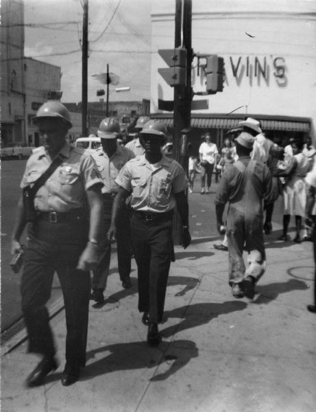 A group of police officers wearing helmets are walking along the sidewalk. Behind them is an intersection, with a number of pedestrians on the sidewalk and crossing the street.