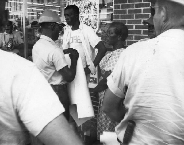 A group of male and female protestors and law enforcement officers stand on the sidewalk outside of a Woolworth's department store during Freedom Summer. One of the officer's is writing on a large sheet of paper. On the far left a man holds a camera.