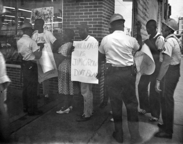 A line of protestors outside of a Woolworth's department store during Freedom Summer hold signs while law enforcement officers stand nearby.  Two of the signs held by the protestors read "Woolworth is Jim Crow Don't Buy" and "[...]Freedom NOW."