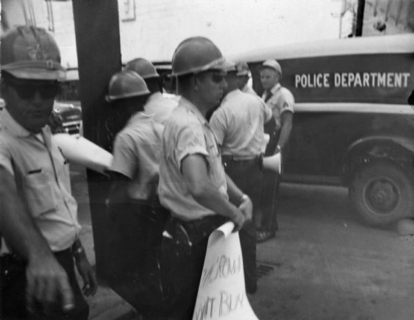 A group of law enforcement officers stand in the street near a vehicle labeled "Police Department" on the side. In the foreground on the left a police officer is pointing at the camera, and next to him another police officer is rolling up a sign that says "[...] Crow [D]on't Buy." The signs were ones that demonstrators had been holding outside of Woolworth's Department store at a protest during Freedom Summer.