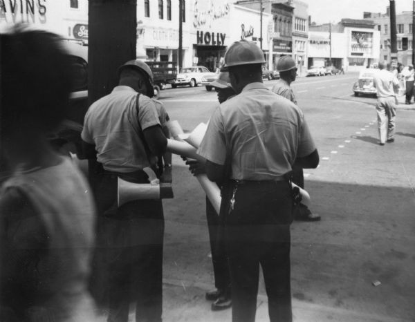 A group of law enforcement officers wearing helmets hold rolled up paper, probably signs confiscated from demonstrators during a Freedom Summer protest outside of Woolworth's department store. In the background, a commercial area is lined with cars and people walking on sidewalks.