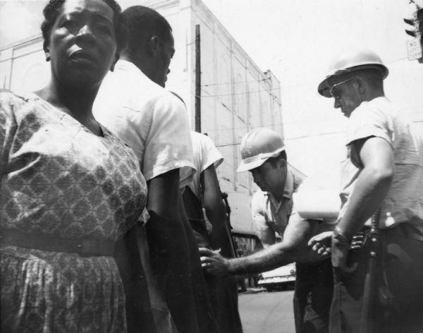 A woman stands in the foreground looking at the camera, while behind her two law enforcement officers wearing helmets examine protestors during a Freedom Summer protest outside a Woolworth's department store.