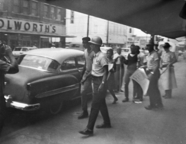 A group of protestors and law enforcement officers walk along the sidewalk across the street from a Woolworth's department store during a Freedom Summer demonstration. Two of the police officers are holding confiscated protest signs.