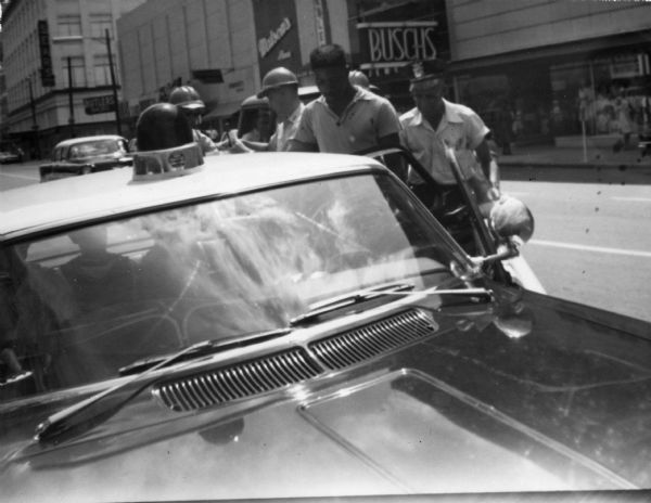 A young man stands at the open passenger door of a police vehicle. A police officer stands directly behind him. A number of passengers are already in the backseat. Three other law enforcement officers wearing helmets are standing in the street behind the squad car.