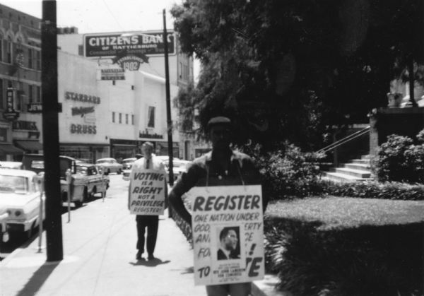 View down sidewalk of two male protestors wearing signs around their necks walking down a sidewalk in a Freedom Summer civil rights demonstration. The signs read "Register / One nation under God [...]" and "Voting is a right not a privilege / Register!"