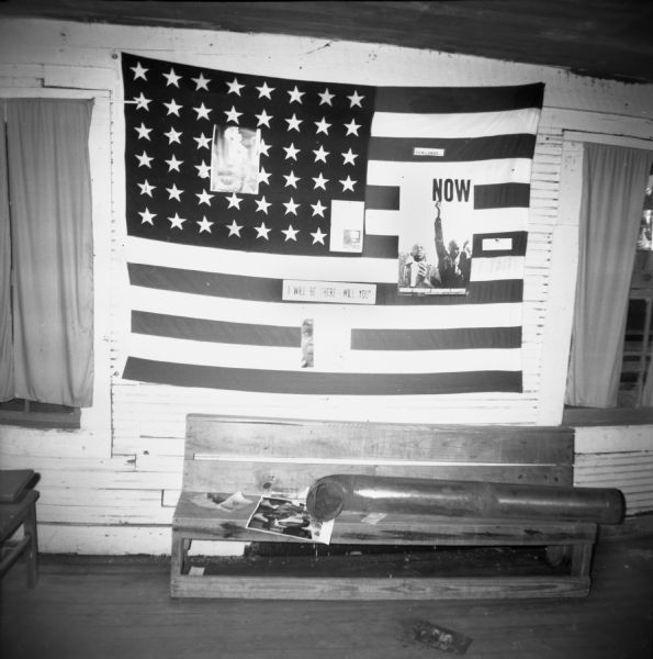 The interior of a Freedom House, used by civil rights workers, is decorated with a United States flag and several posters on a wall. A bench below the flag has what appears to be a large stove pipe, as well as papers on it.