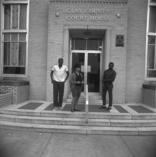Two men and a woman stand outside the entrance to the Clay County Courthouse. To the right of the doorway is a sign that reads "Fallout Shelter." The caption on the back of the image reports that "on the bulletin board inside was an advertisement for the Klan."