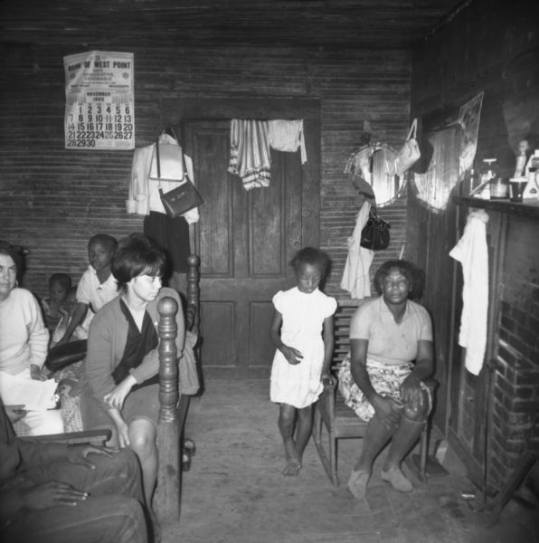 Three women, another adult seated in a chair on the left, and three children are gathered in a small room. "Mrs. Lester Davidson, candidate for the ASCS (Agricultural Stabilization and Conservation Service) talks to MFDP (Mississippi Freedom Democratic Party) workers in one of her two rooms. Nine people live there."